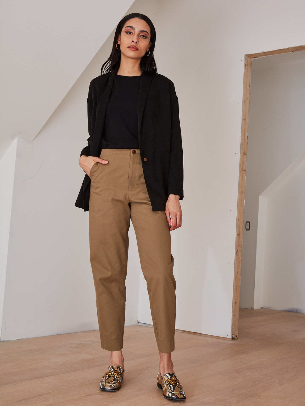 A woman stands in a minimalist room with white walls and light wood flooring, wearing a black blazer over a black top, tan cropped trousers, and patterned loafers.