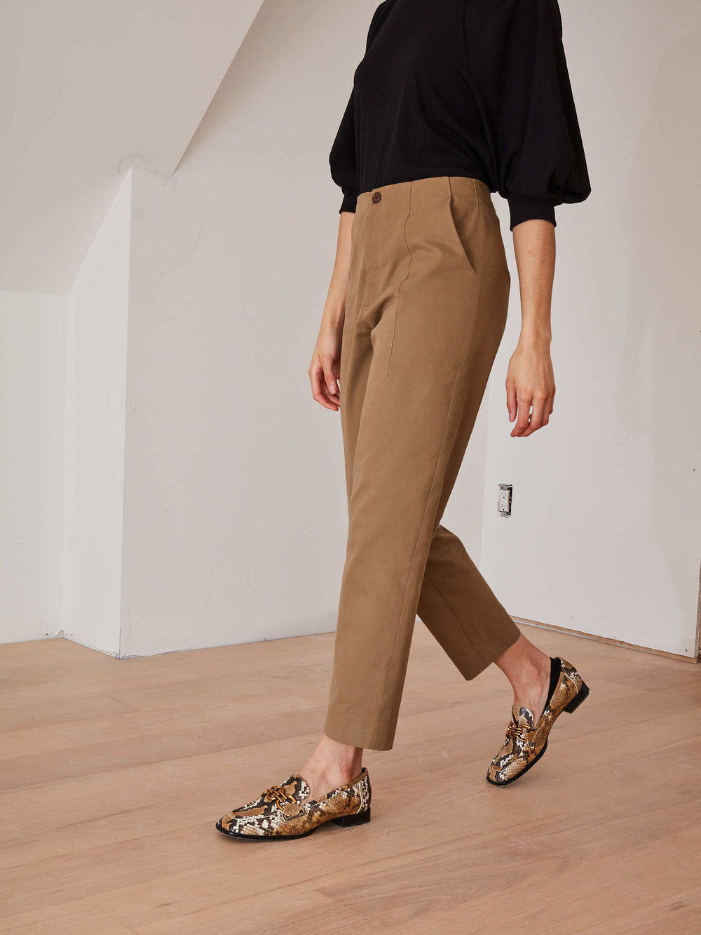 A model wearing high-waisted, straight-leg brown trousers, a black top, and brown and gold patterned loafers, walking in a minimalist room with white walls and a light wood floor.