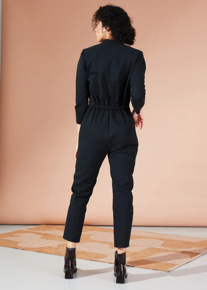 A model viewed from behind wearing a black long-sleeve jumpsuit with a gathered elastic waist and slim cropped legs, paired with black ankle boots, standing on a patterned rug against a peach studio backdrop.
