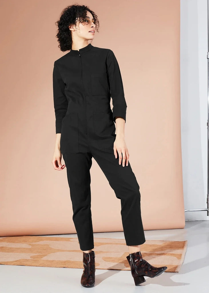 A model wearing a black long-sleeve jumpsuit with a mock neck and zippered front, paired with dark brown ankle boots, standing against a peach-colored studio backdrop.