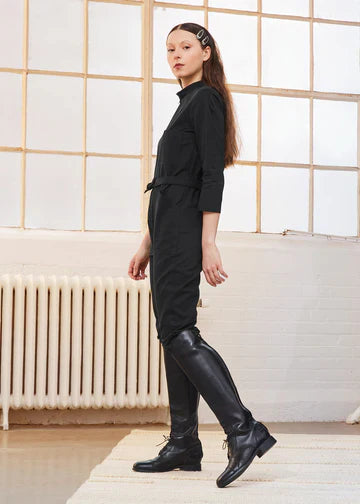 A woman modeling a black long-sleeve jumpsuit with a waist tie, paired with black leather riding boots, standing in a bright room with a large window and radiator.