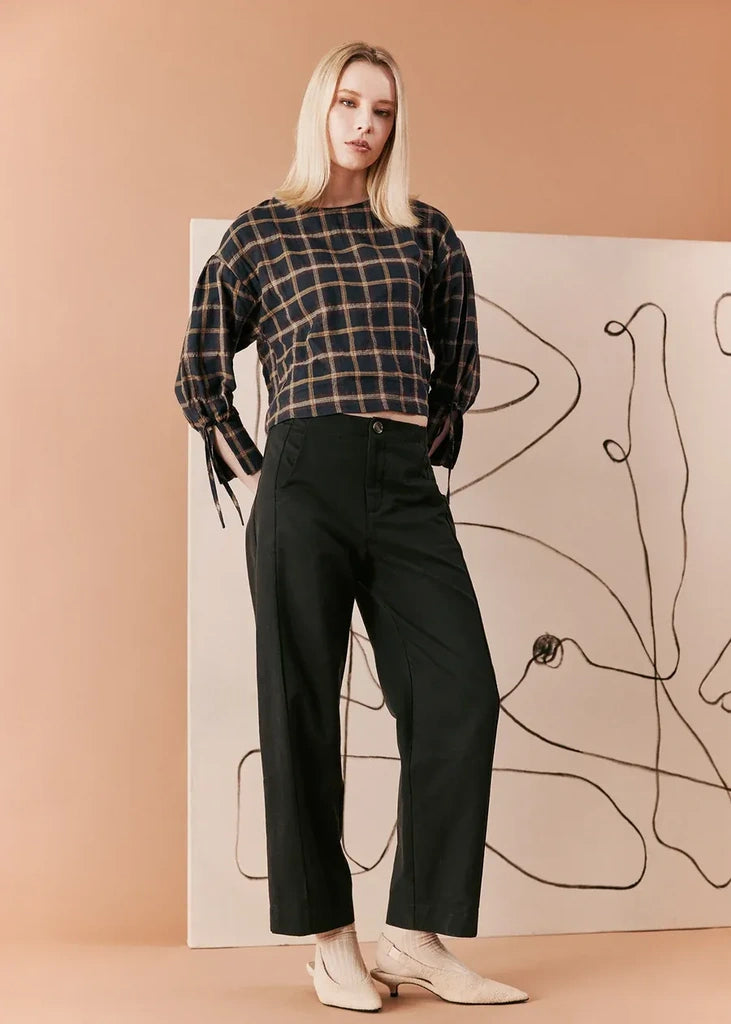 A blonde model stands in a studio wearing a dark plaid cropped long-sleeve top and black wide-leg cropped pants, paired with off-white pointed-toe heels. She is posed in front of a large canvas print with abstract black line art against a warm peach background.