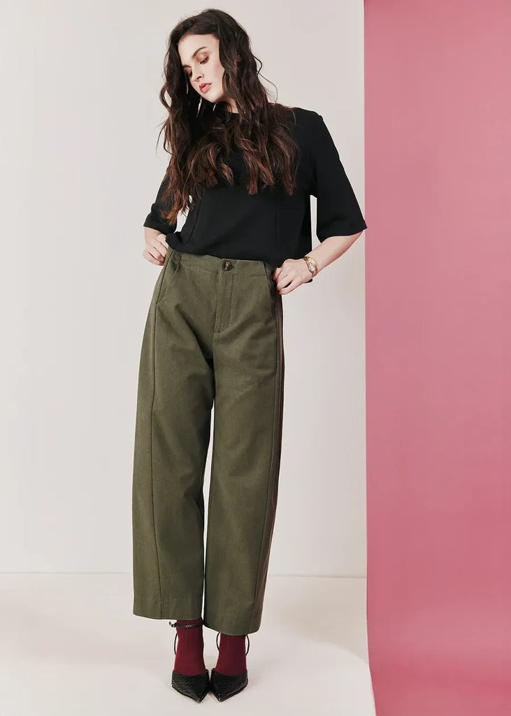 A woman modeling a pair of olive green wide-leg cropped trousers, styled with a black short-sleeve top, burgundy ankle socks, and black pointed-toe heels, standing against a split white and pink studio background.