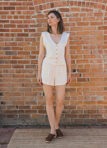 A woman wearing a sleeveless cream-colored romper with a button front and matching shorts, standing against a red-brown brick wall.