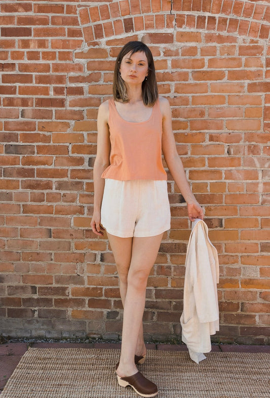 A woman stands against a red brick wall wearing a peach tank top, off-white shorts, and brown clogs, while holding a light beige jacket.