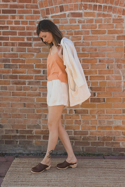 A woman wearing a peach tank top, white shorts, and brown clogs walks past a red brick wall while carrying a white jacket over her shoulder.