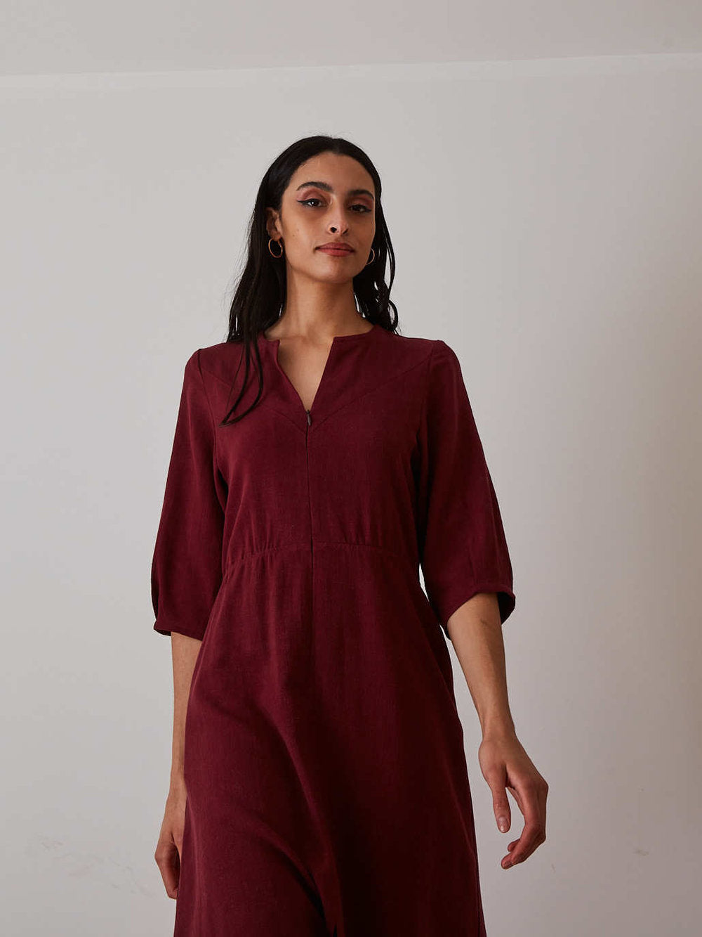 A woman modeling a dark burgundy midi dress with three-quarter sleeves and a split V-neckline, standing against a plain off-white wall.