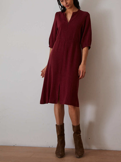 A woman wearing a knee-length burgundy midi dress with three-quarter sleeves and a split neckline, paired with brown suede ankle boots, standing against a plain off-white wall.