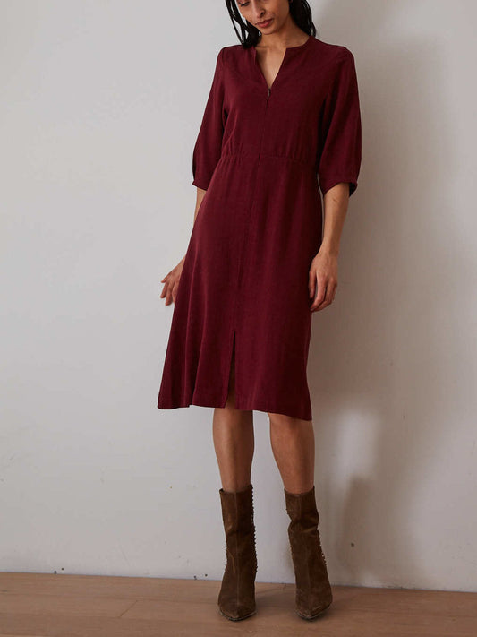A woman wearing a knee-length burgundy midi dress with three-quarter sleeves and a split neckline, paired with brown suede ankle boots, standing against a plain off-white wall.