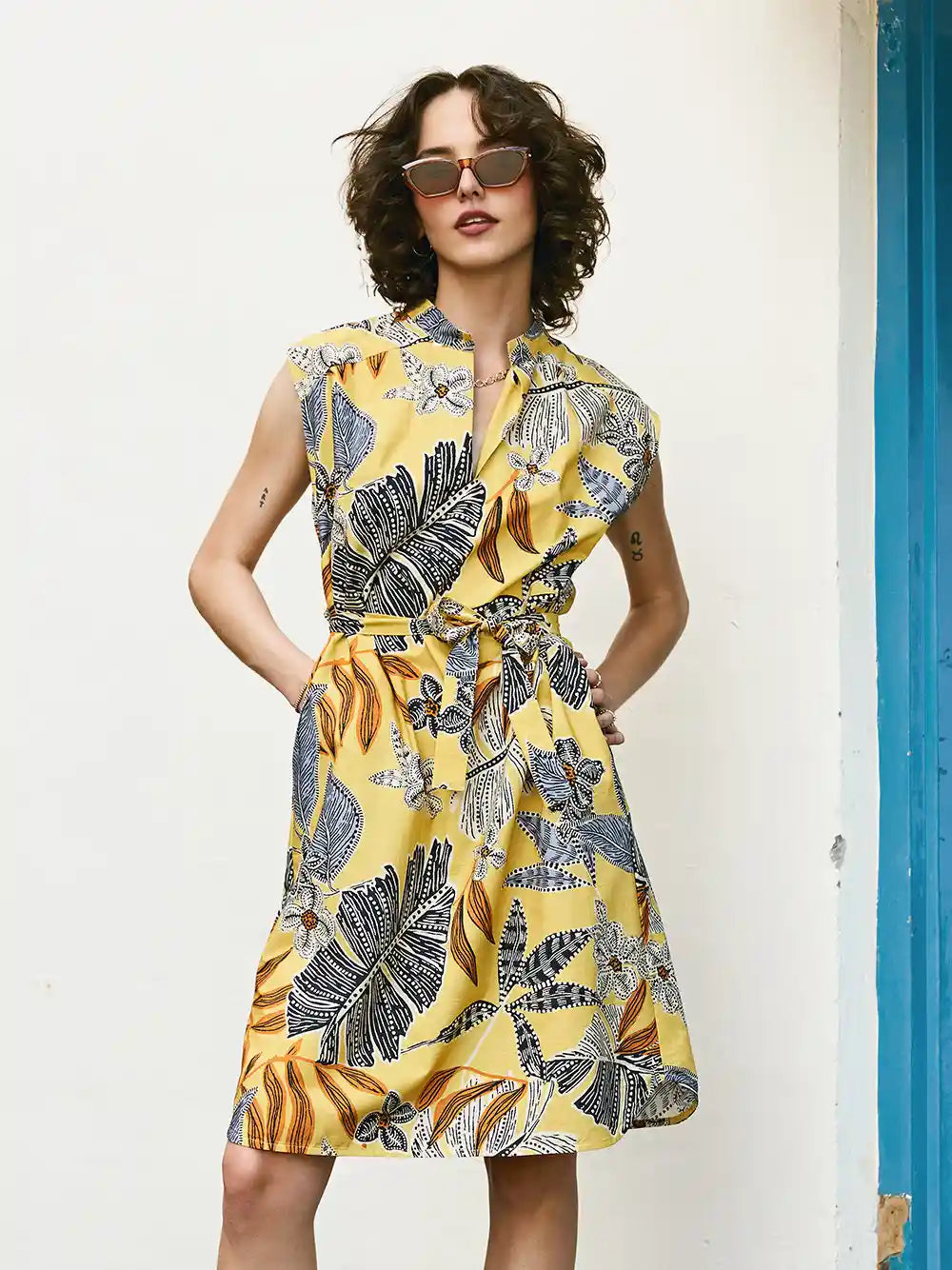 Woman wearing a yellow floral dress against a white wall.