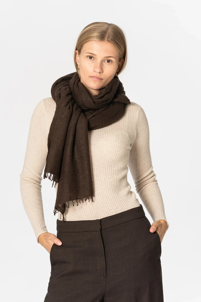 A woman modeling a dark brown scarf wrapped around her neck, paired with a beige ribbed long-sleeve top and dark brown trousers, against a light gray studio background.
