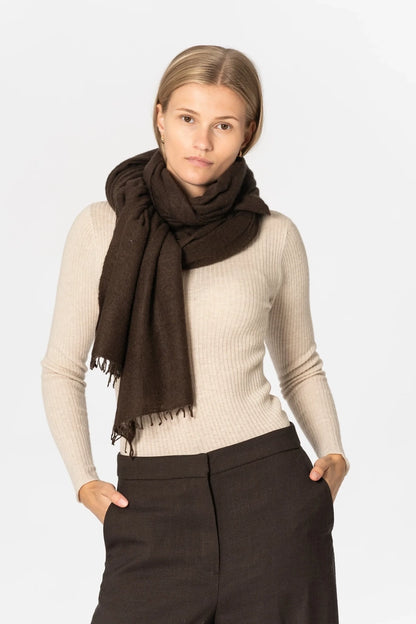 A woman modeling a dark brown scarf wrapped around her neck, paired with a beige ribbed long-sleeve top and dark brown trousers, against a light gray studio background.