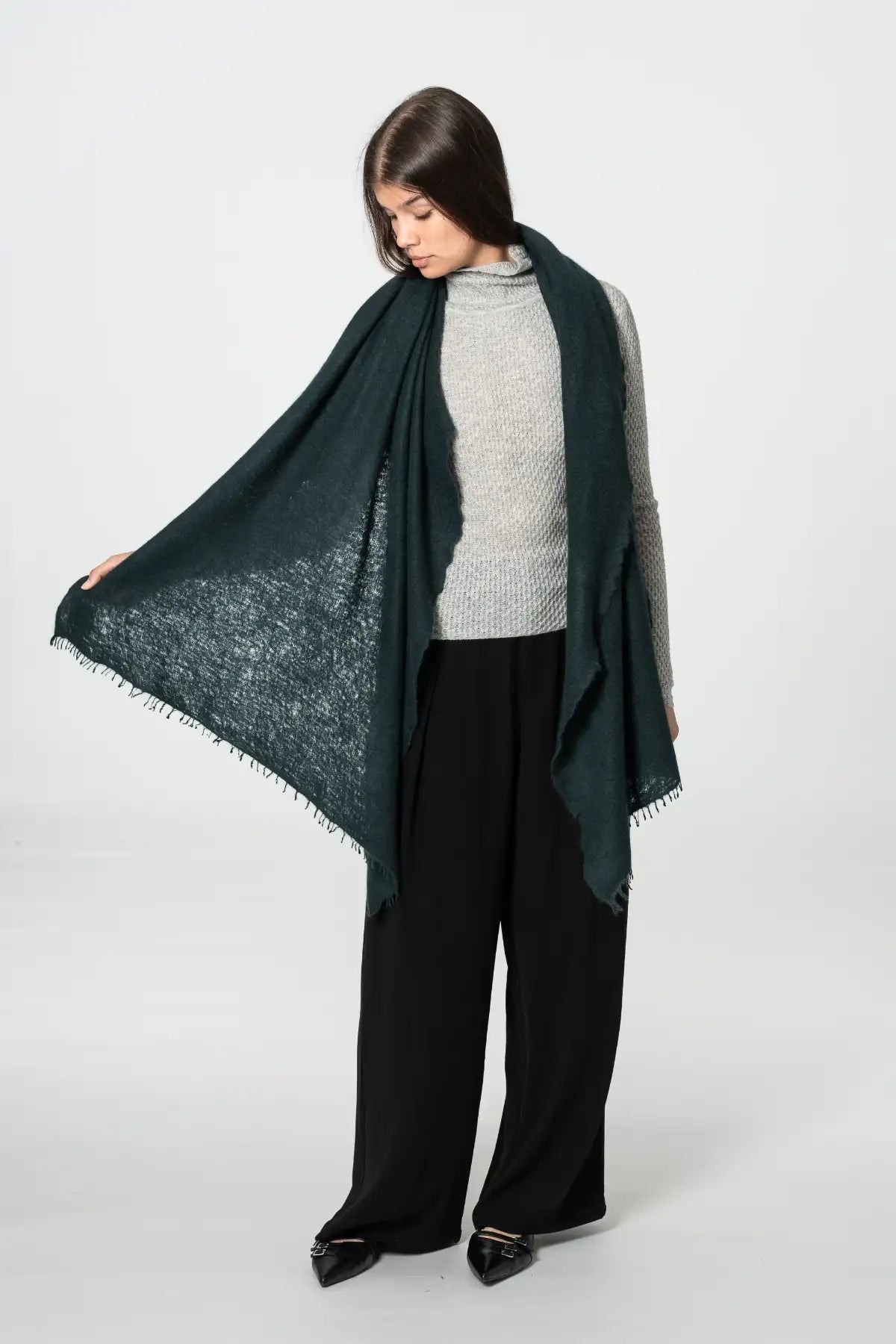A woman modeling a dark peacock green scarf with a textured openwork pattern and fringed edges, draped over her shoulders and held out to the side against a light gray studio background.
