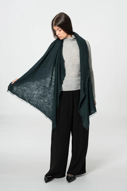 A woman modeling a dark peacock green scarf with a textured openwork pattern and fringed edges, draped over her shoulders and held out to the side against a light gray studio background.