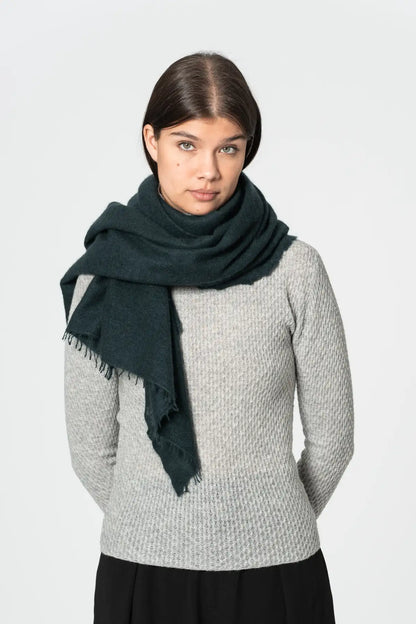 A model wearing a dark peacock green scarf with fringed edges over a light gray ribbed knit sweater and a black skirt, photographed against a solid light gray background.