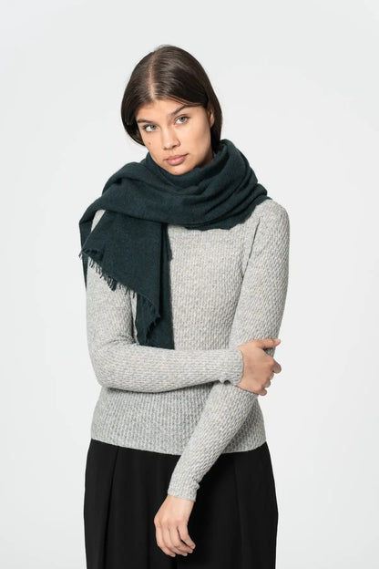 A woman modeling a dark peacock green fringed scarf wrapped around her neck, paired with a light gray knit sweater and black skirt, against a plain light gray background.