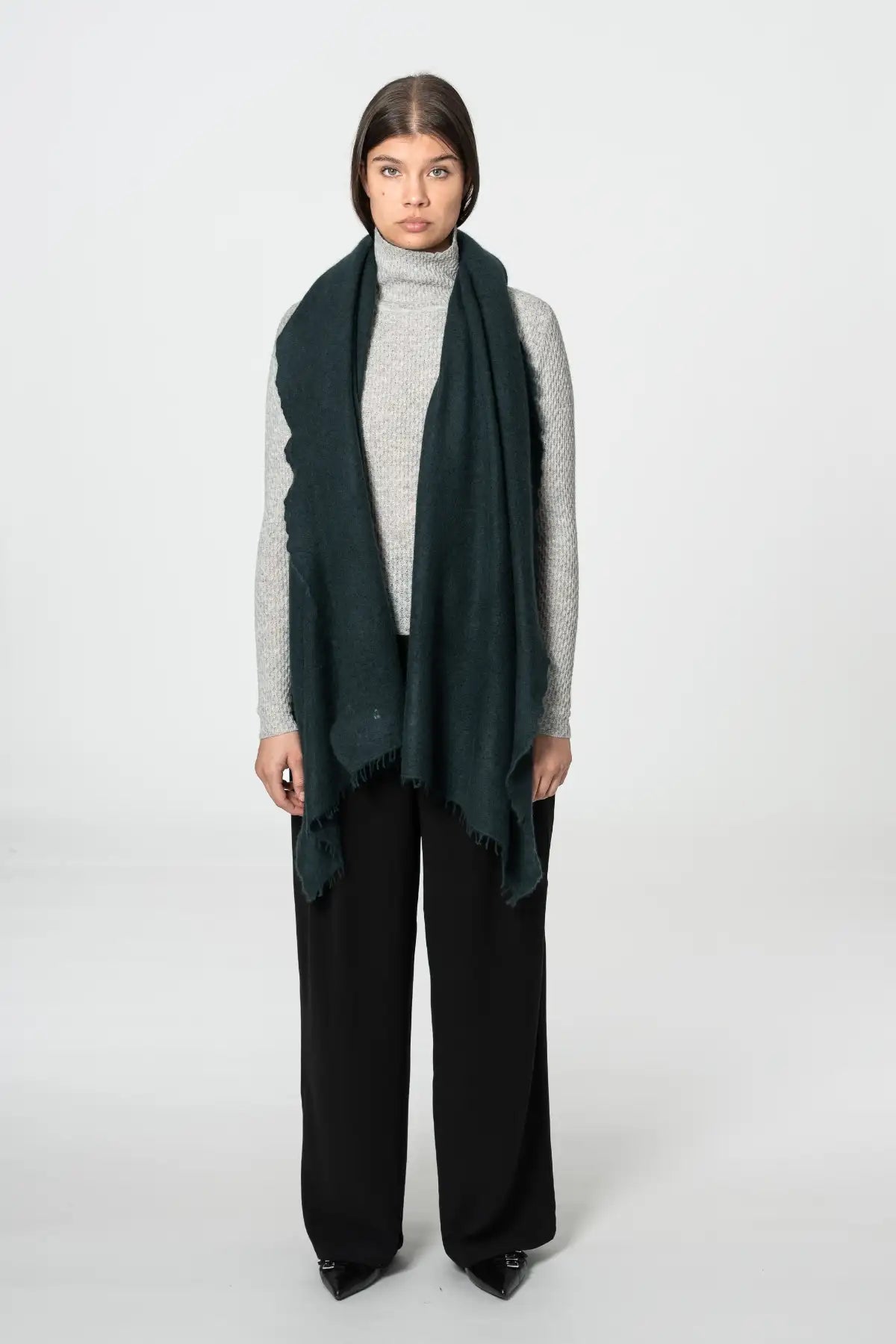 A model stands against a light gray studio background wearing a dark peacock green scarf draped over a light gray turtleneck sweater and black wide-leg trousers.