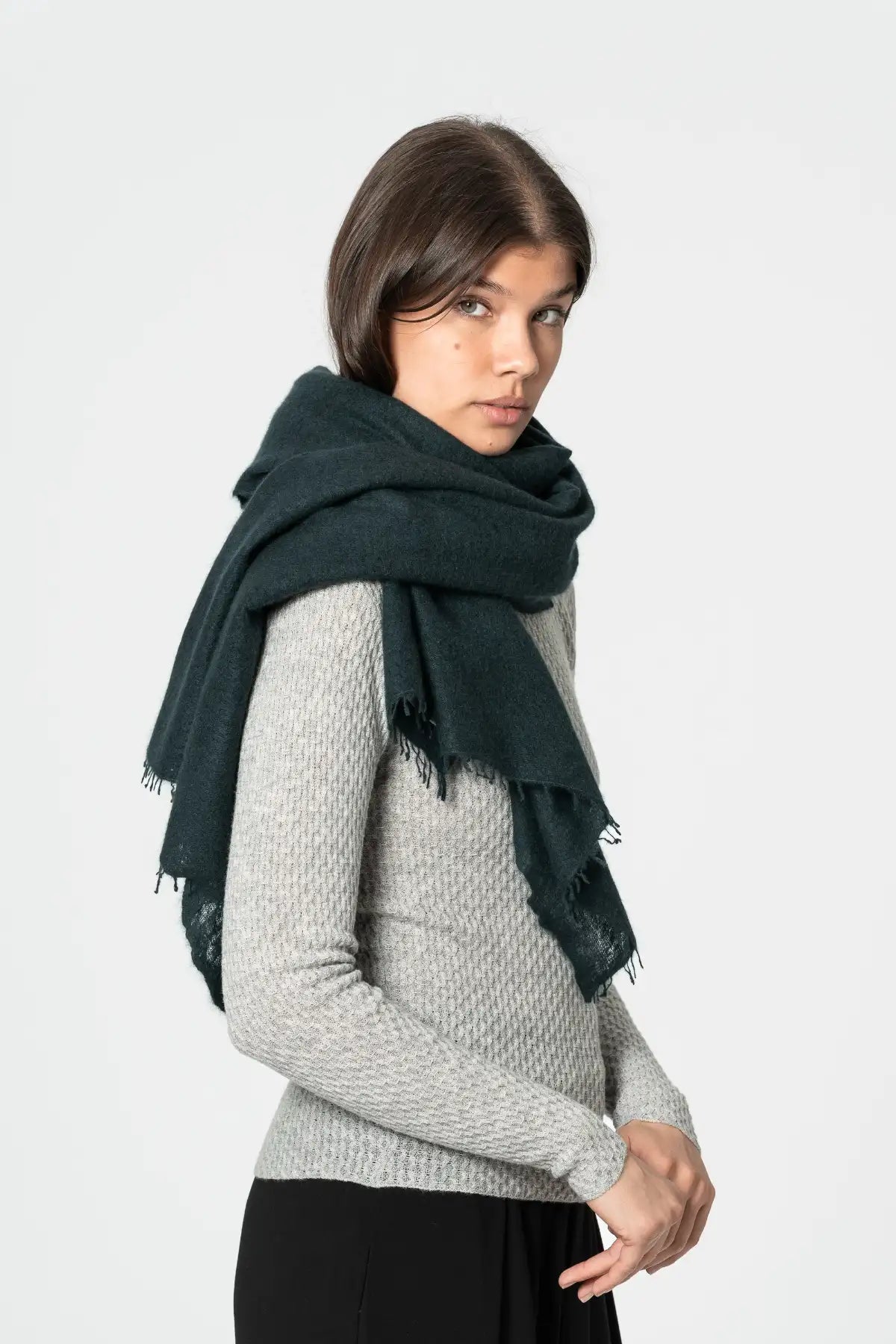 A woman wearing a dark peacock green scarf with fringed edges over a light gray knit sweater and black pants, photographed against a plain light gray studio background.