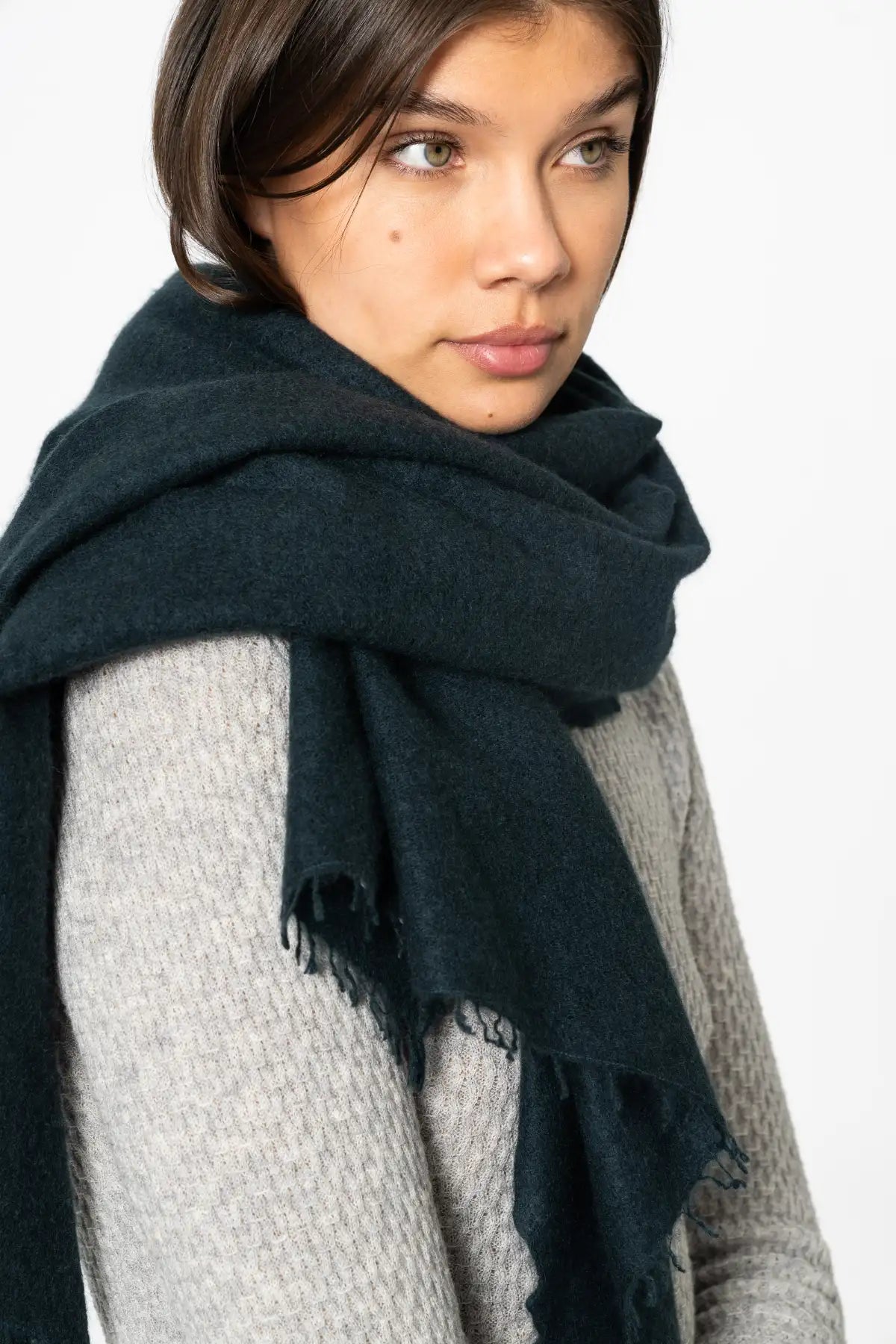 A close-up of a model wearing a dark peacock green fringed scarf wrapped around her neck over a light gray knit sweater, set against a plain white background.