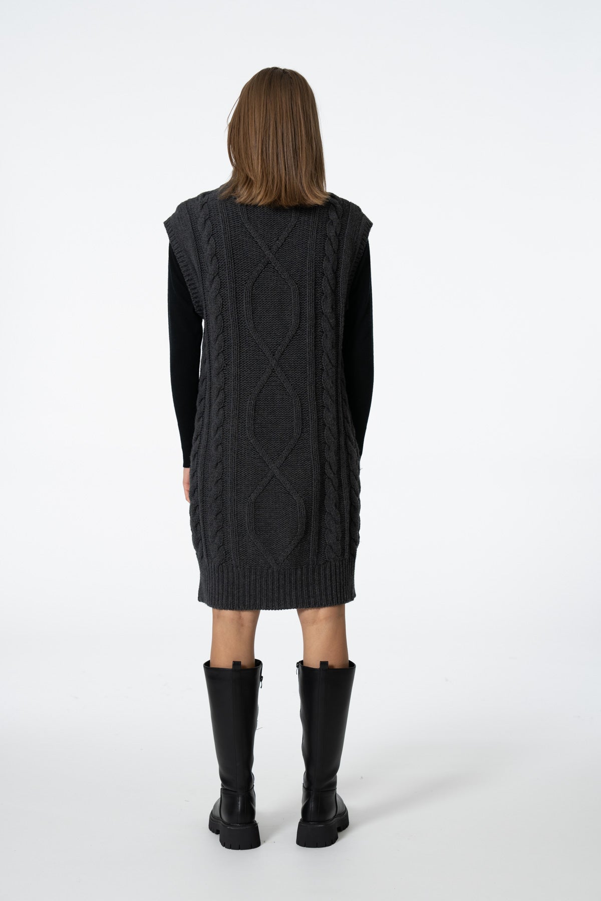 A model viewed from the back wearing a dark charcoal gray cable-knit sweater vest over a black long-sleeve top, paired with black knee-high boots, against a plain white studio background.