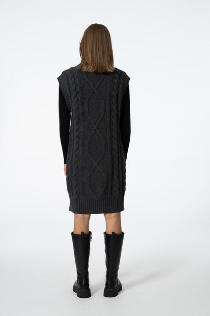 A model viewed from the back wearing a dark charcoal gray cable-knit sweater vest over a black long-sleeve top, paired with black knee-high boots, against a plain white studio background.