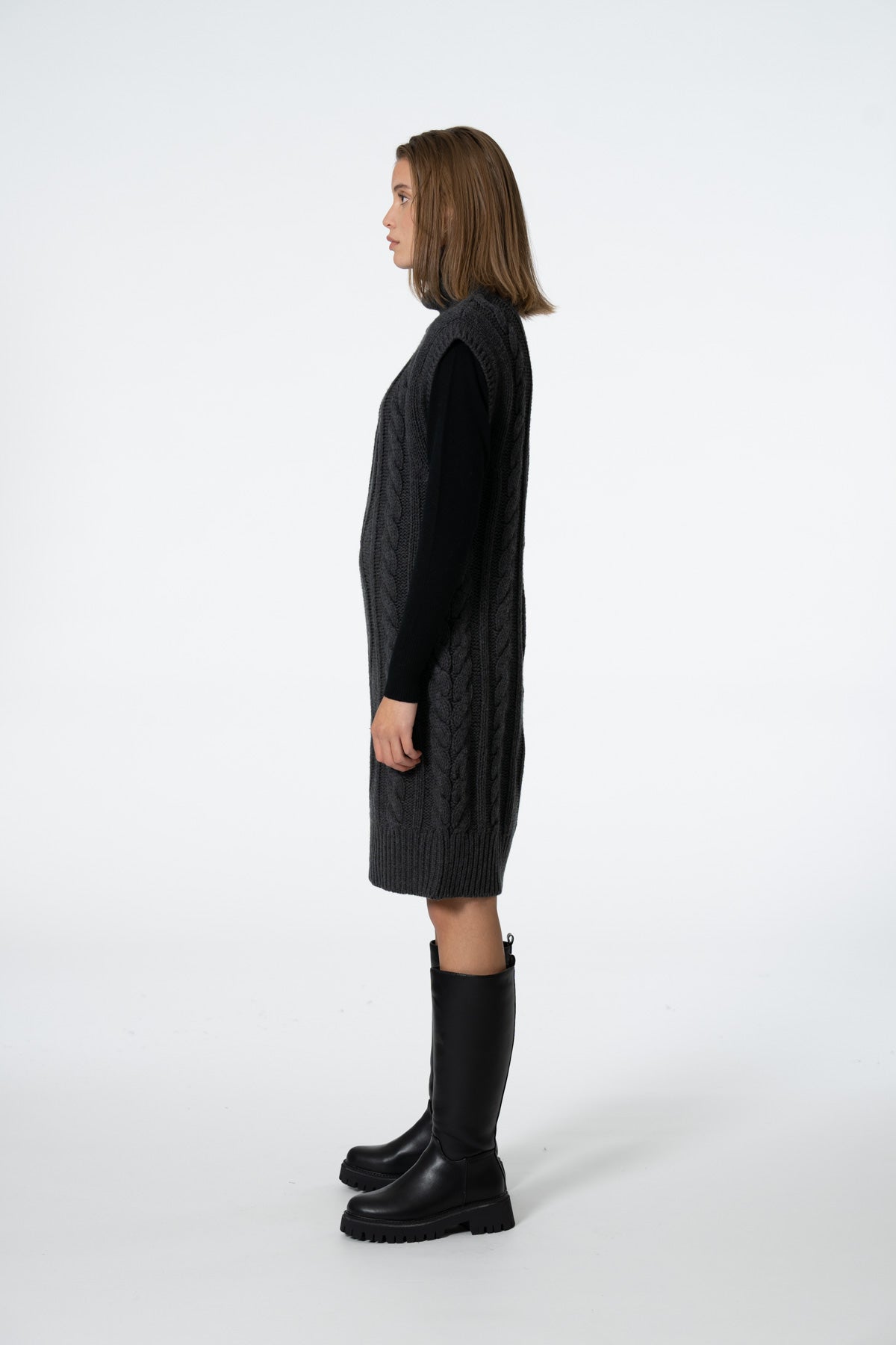 A woman shown in profile wearing a black cable-knit sleeveless sweater vest over a black long-sleeve top, paired with black knee-high boots, against a plain white studio background.