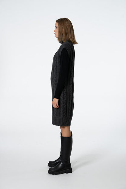A woman shown in profile wearing a black cable-knit sleeveless sweater vest over a black long-sleeve top, paired with black knee-high boots, against a plain white studio background.