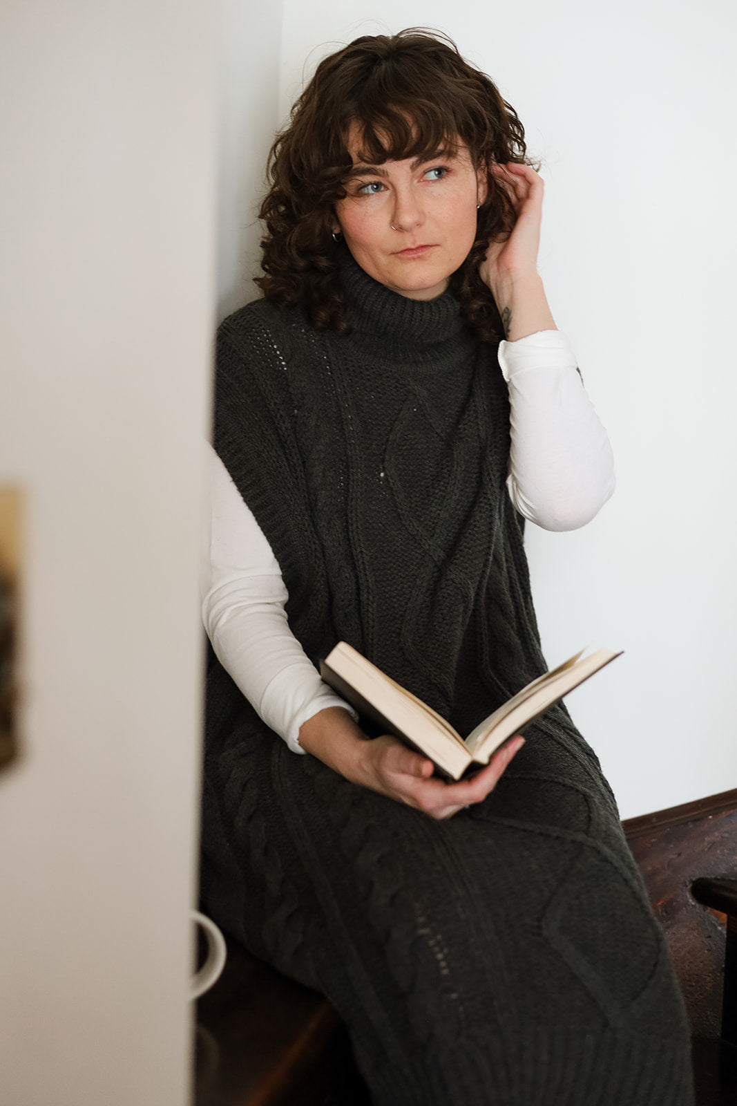 A woman with curly brown hair sits in a white room, wearing a dark gray cable-knit turtleneck sweater dress over a white long-sleeve shirt, and holding an open book.