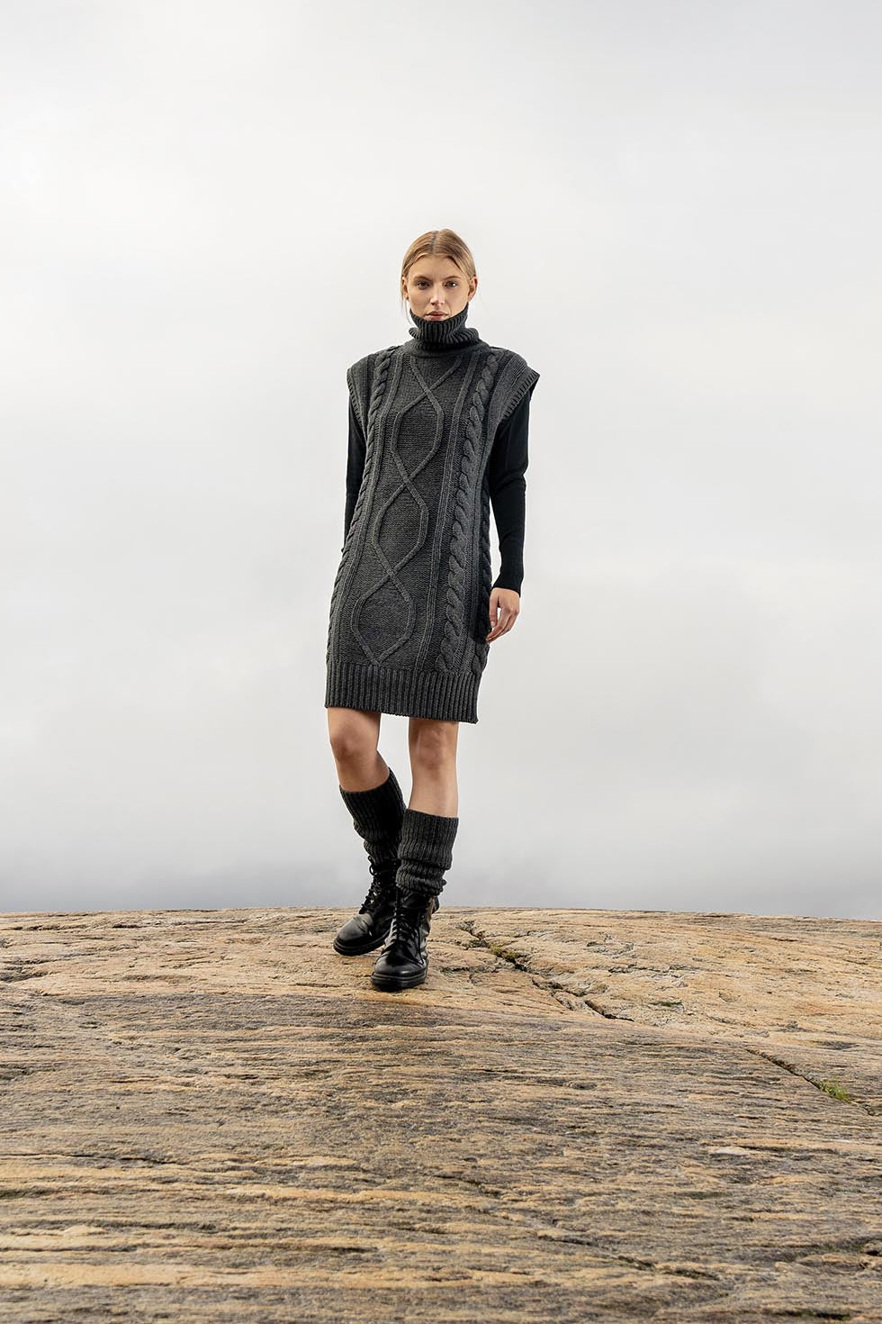 A model stands on a rocky outcrop wearing a dark gray cable-knit sleeveless turtleneck dress over a black long-sleeve top, paired with black knee-high socks and black boots, against a misty gray sky.