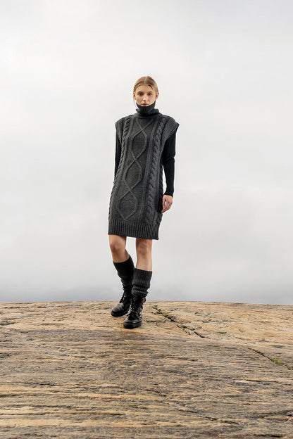 A model stands on a rocky outcrop wearing a dark gray cable-knit sleeveless turtleneck dress over a black long-sleeve top, paired with black knee-high socks and black boots, against a misty gray sky.