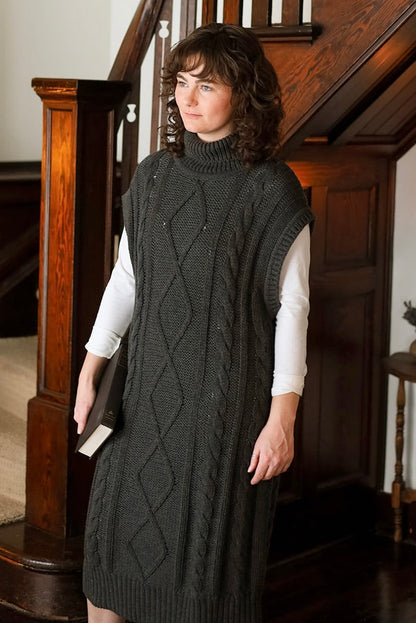 A woman wearing a dark charcoal cable-knit mock-neck sweater dress over a white long-sleeve shirt, standing in a home interior near a wooden staircase.