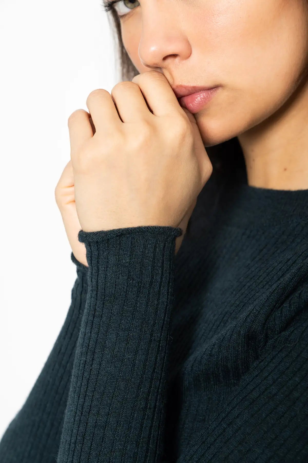 Close-up of a person wearing a dark ribbed knit sweater, with the hands resting near the mouth against a plain white background.
