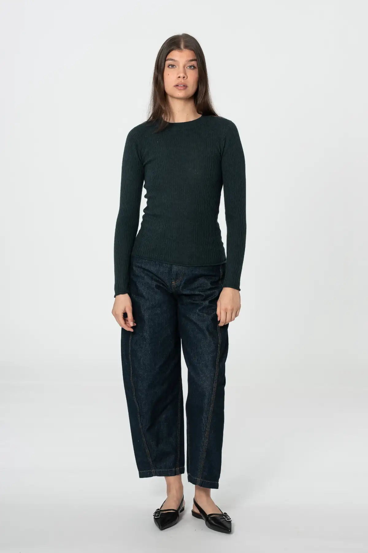 A woman stands against a light gray studio background wearing a dark green long-sleeve knit top, dark blue cropped jeans, and black pointed flats.
