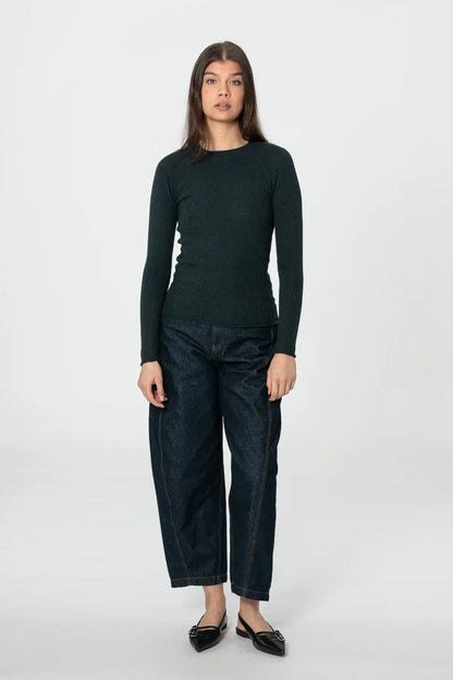 A woman stands against a light gray studio background wearing a dark green long-sleeve knit top, dark blue cropped jeans, and black pointed flats.