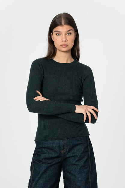 A woman modeling a dark forest green ribbed knit crewneck sweater with dark blue jeans, standing against a plain light gray background.