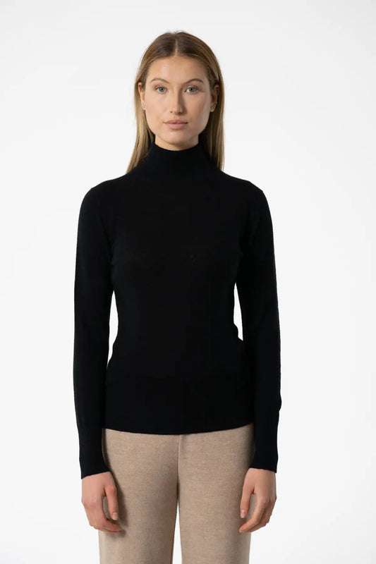 Woman wearing the Dinadi Merino Turtleneck in Black, a slim fit extra fine merino wool knit with a tall turtleneck and long sleeves, tucked into oatmeal knit trousers against a white studio background.