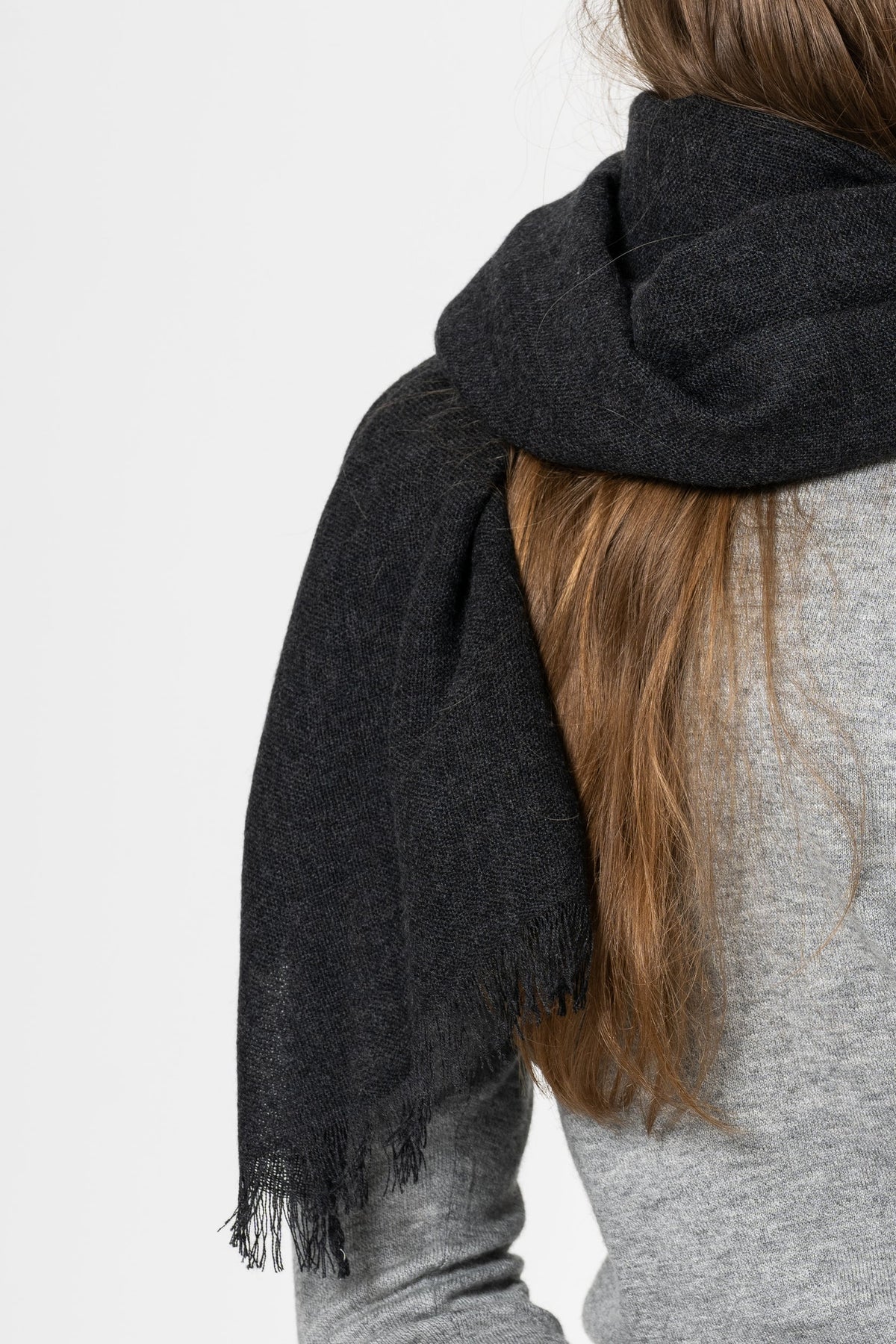 A close-up of a person wearing a dark charcoal gray fringed scarf over a light gray top, shown from the back against a plain white background.