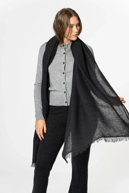 A woman modeling a lightweight black scarf with fringed edges, draped over her shoulders over a gray cardigan and black pants, against a plain white studio background.
