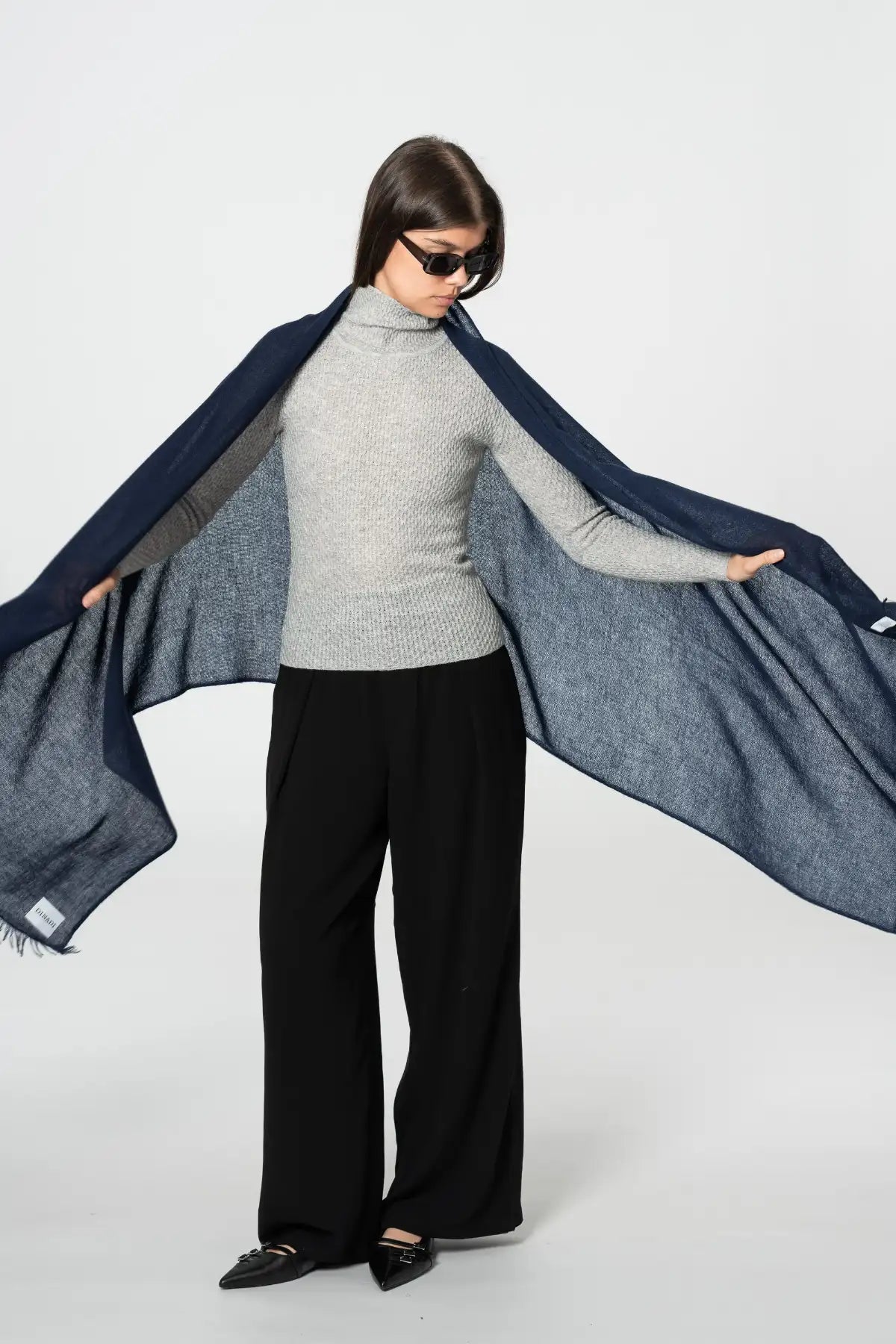 Woman wearing a gray sweater, black pants, and sunglasses, holding a blue scarf.