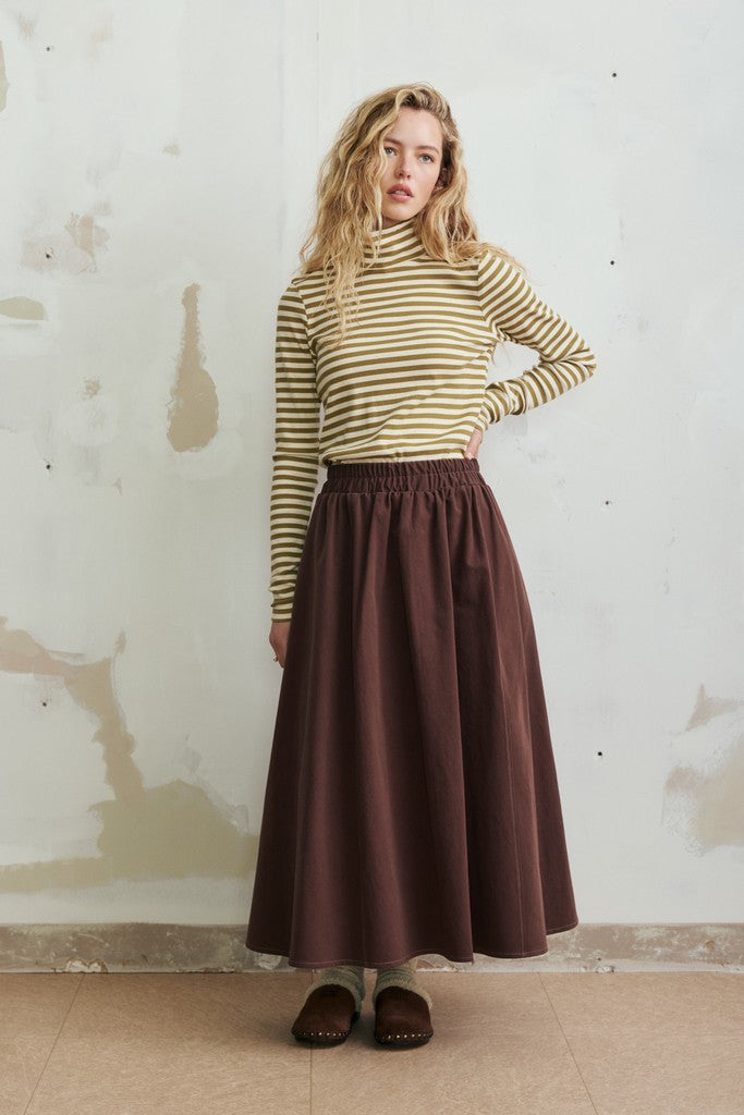A woman stands against a textured off-white wall wearing a mustard yellow and white striped mock-neck long-sleeve top and a dark brown pleated maxi skirt, paired with dark brown slip-on shoes.