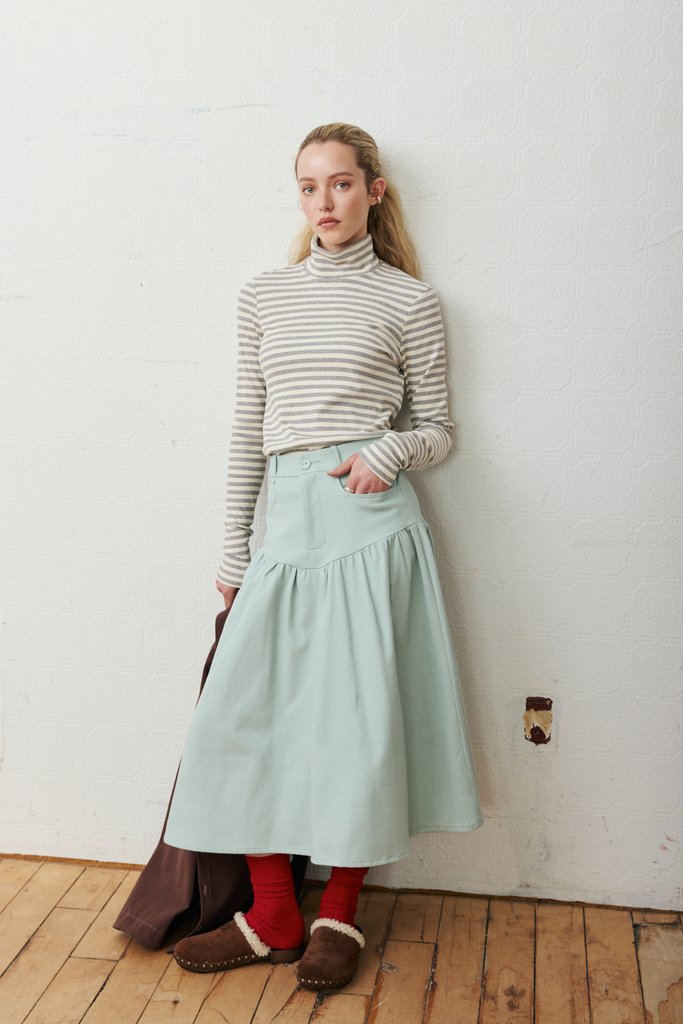 A woman stands against a white wall wearing a beige and white striped turtleneck sweater, a light mint green tiered skirt, red socks, and brown slippers. She is holding a dark brown bag on a light wood floor.
