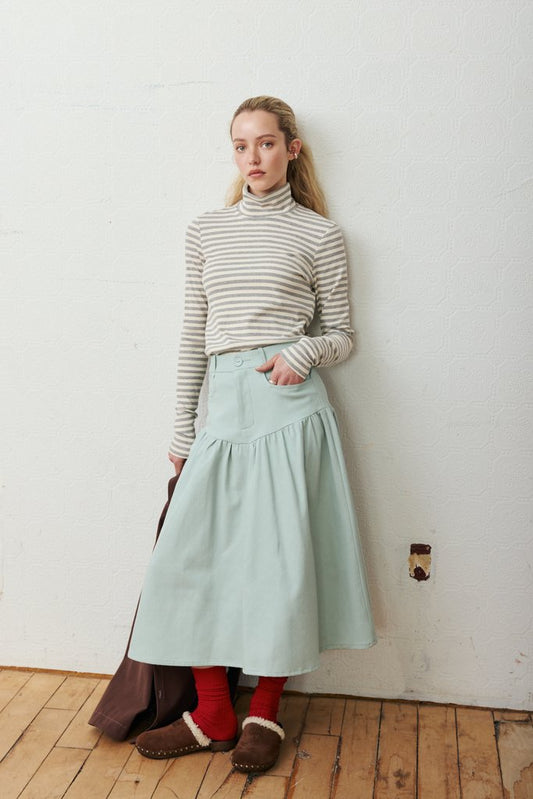 A woman stands against a white wall wearing a beige and white striped turtleneck sweater, a light mint green tiered skirt, red socks, and brown slippers. She is holding a dark brown bag on a light wood floor.