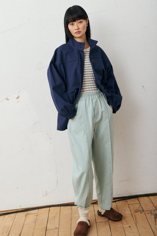 A model stands against a white wall on a wooden floor, wearing a navy blue jacket over a striped shirt and light sage green pants.