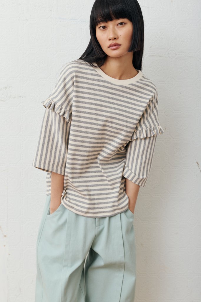 A woman with a straight black bob haircut wearing a beige and gray striped ruffle-sleeve top and light sage green trousers, standing against a white textured wall.
