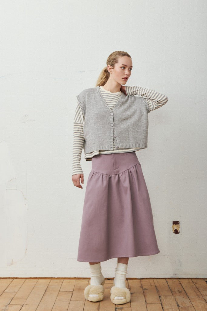 A woman stands in a room with a white wall and wooden floor, wearing a light gray knit cardigan over a cream and gray striped long-sleeve shirt, a dusty purple pleated midi skirt, white socks, and fluffy off-white slip-on shoes.