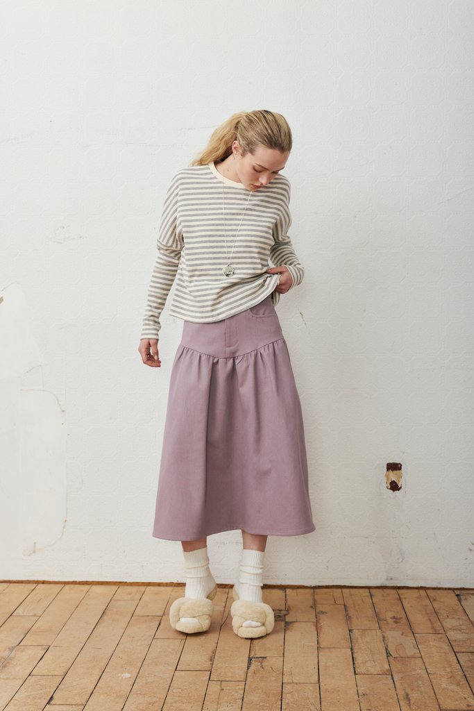 A woman stands in a room with a white wall and wooden floor, wearing a beige and gray striped long-sleeve top, a dusty purple midi skirt, white socks, and beige slippers.