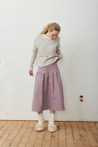 A woman stands in a room with a white wall and wooden floor, wearing a beige and gray striped long-sleeve top, a dusty purple midi skirt, white socks, and beige slippers.