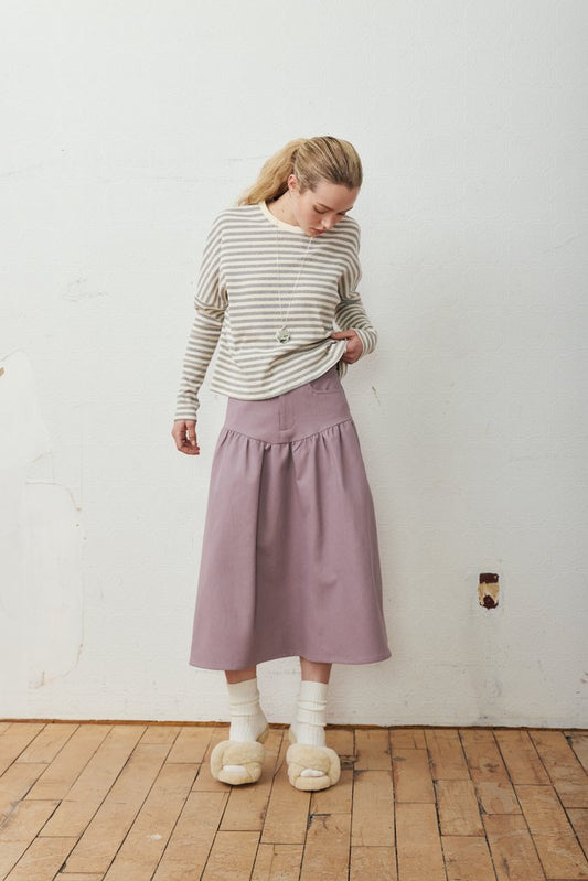 A woman stands in a room with a white wall and wooden floor, wearing a beige and gray striped long-sleeve top, a dusty purple midi skirt, white socks, and beige slippers.