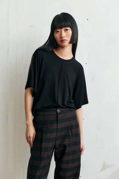 A model with a straight black haircut stands against a light off-white wall, wearing a black short-sleeve top and dark brown and black horizontally striped trousers.