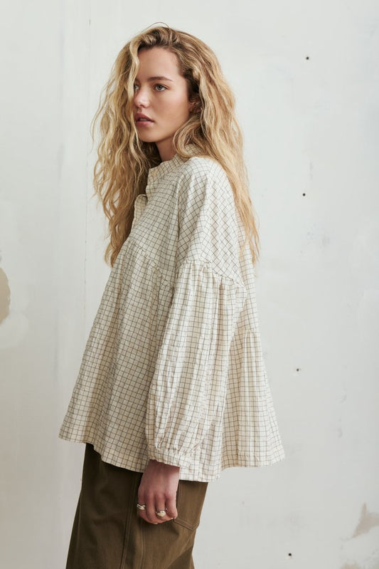 A woman with long wavy blonde hair models a cream and tan gingham long-sleeve blouse with a high collar and voluminous sleeves, paired with olive brown pants, standing against a textured off-white wall.